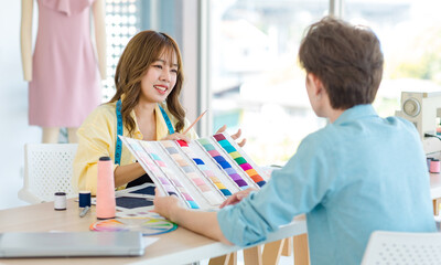 Millennial Asian young cheerful professional female dressmaker designer seamstress with measuring tape sitting smiling discussing choosing color with unrecognizable male customer in studio office