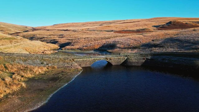 Winter aerial footage of a small stone built bridge crossing a reservoir and river high on the Pennine hills, West Yorkshire, England. With blue water and frosty fields.