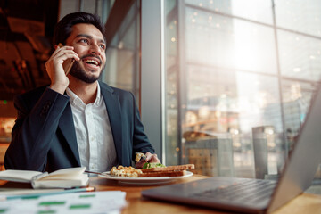 Smiling indian businessman is talking phone with client during lunch time in cozy cafe