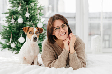 Image of attractive woman with gentle smile, has dark hair, wears transparent glasses spends time with lovely pet, lie on white bed against New Year tree. Merry Christmas and happy winter holidays