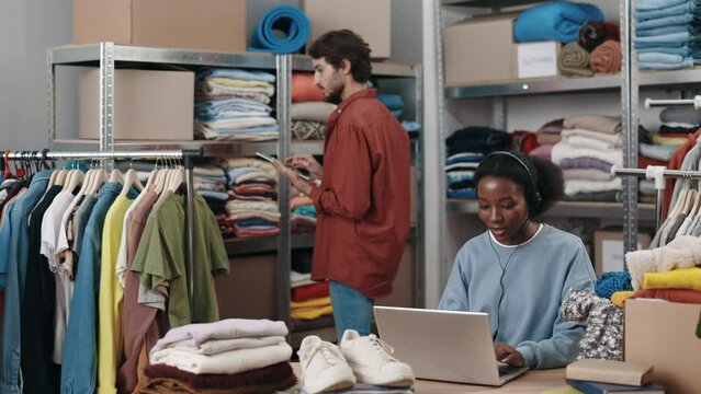 Multiracial woman wearing headphones sitting at the laptop and talking with client while working at the second hand or reuse clothes center. Sustainability and call center concept