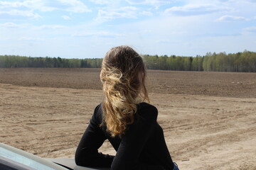 a girl with long blonde hair that flutters in the wind is leaning on a car and looking out into the field