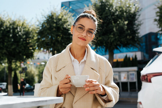 Cute Stylish Young Mixed Race Woman Relaxing In Street Cafe Holding Coffee Mug Looking At Camera Outdoors. Portrait Of Brunette Woman Wearing Glasses Having Breakfast In Street Cafe On Sunny Day