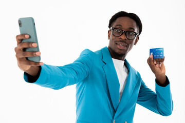 African american young guy posing on white background in studio holding credit card in ahnds and cellphone talking by videolink with girlfriend in blue elegant suit.