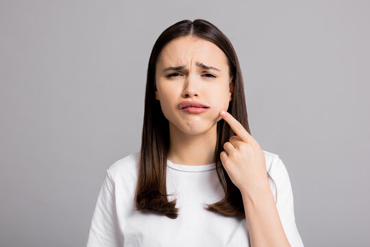 Crying tired sad woman suffers from painful toothache dental illness pointing on teeth standing on grey background in studio isolated wearing basic white t-shirt.