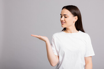 Delighted adorable beautiful woman in white casual t-shirt standing on grey copyspac background opening hands palm up like hoding something on.