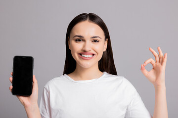 Portrait close up shot on young woman girl in basic white t-shirt standing on grey background in studio isolated holding new last series smartphone selling something on black screen blank shows okay.