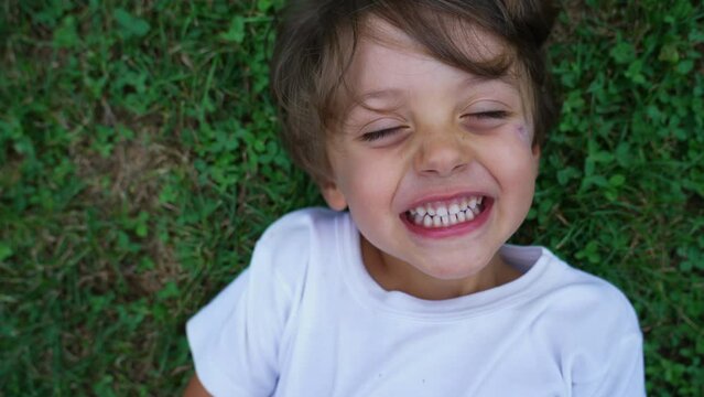 Happy child laughing close up face. Male kid laid on grass smiling. Real life laugh and smile. Authentic joy expression