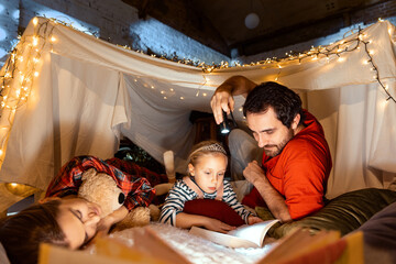 Obraz na płótnie Canvas Happy mother, father and daughter lying inside self-made hut, tent in room and reading tales. Cozy family evening