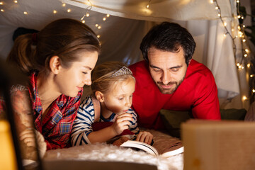 Happy family, mother, father and daughter in pajamas lying inside self-made hut, tent in room in the evening and reading stories