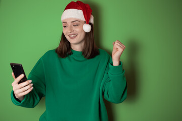 Cheerful young woman dressed in a green sweater and santa hat, holding a mobile phone in her hand, making a winner gesture, isolated on a green background. Happy new year 2023 holiday concept