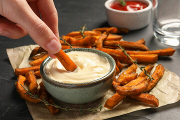 Woman dipping sweet potato fries into sauce at dark grey table, closeup - Powered by Adobe