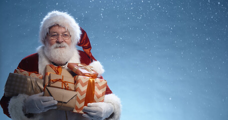 Close up shot of senior bearded man in santa clause outfit carrying gift boxes, isolated on blue background - christmas spirit, holiday mood 