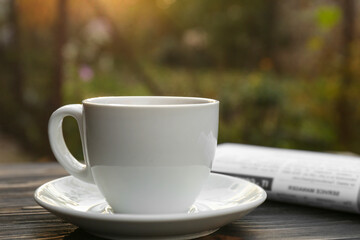 White cup with coffee and newspaper on wooden table, closeup. Morning ritual