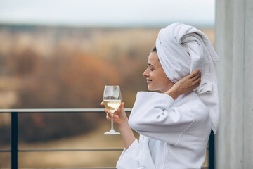 A girl in a white bathrobe and a glass of wine enjoys relaxation and spa treatments - obrazy, fototapety, plakaty