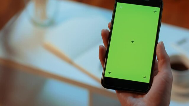 (Camera used: ARRI ALEXA, 50mm lens, slow motion) Slow pan of a Caucasian hand holding a green screen cell phone with tracking marks, with a cup of coffee in the background.
