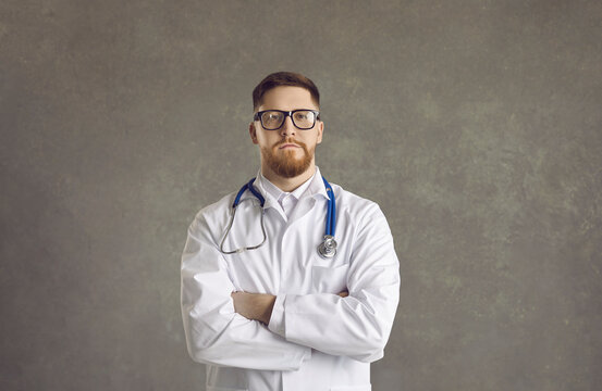 Serious Confident Male Doctor Wearing Lab Coat And Stethoscope Standing With Crossed Hand On Chest And Looking At Camera Portrait Studio. Adult General Practitioner In Glasses. Headshot Medical Staff