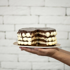 Cake in cut form on platter in hands. Festive chocolate dessert with buttercream for home celebration. Background of white brickwork. Soft focus. Side view. Sectional view.