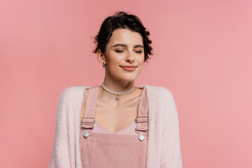 pleased woman in strap dress and cardigan smiling with closed eyes isolated on pink