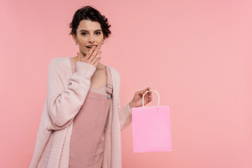 Fototapeta na wymiar amazed woman in warm cardigan holding shopping bag and covering open mouth with hand isolated on pink