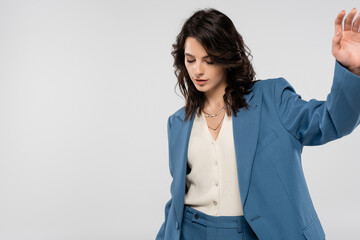 trendy brunette woman in blue blazer standing with outstretched hand isolated on grey