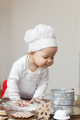 An Asian baby in a chef's hat and apron is preparing Christmas ginger cookies. With curiosity, he looks into the container with flour