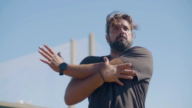 Strong Hispanic man stretching arms, working out outdoors. Male athlete training outdoors on windy summer day alone, looking determined and powerful. Motivation, strength, sport concept.