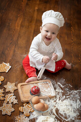 An Asian baby in a chef's cap and apron sitting on the kitchen table and smeared in flour prepares Christmas ginger cookies