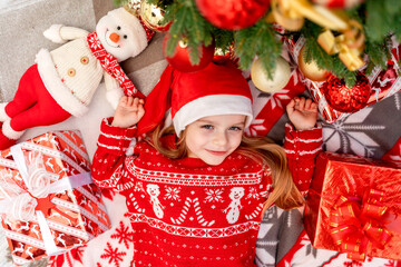 a happy baby girl lies under the Christmas tree at home and waits for the new year or Christmas in a red Santa Claus hat and smiles and rejoices