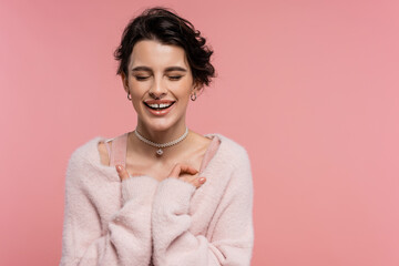 cheerful woman in fluffy cardigan and pearl necklace laughing with closed eyes isolated on pink