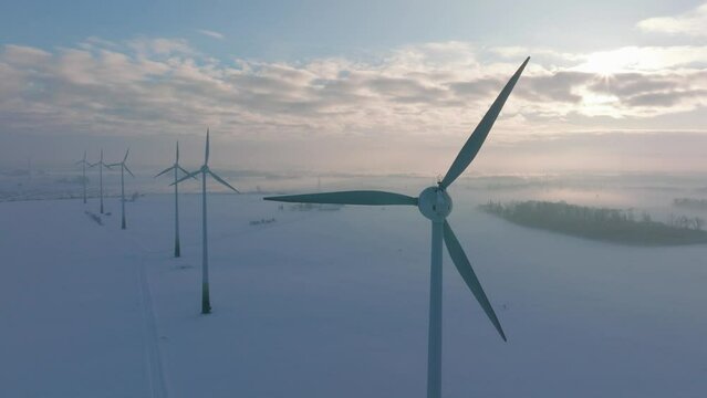 Aerial establishing view of wind turbines generating renewable energy in the wind farm, snow filled countryside landscape with fog, sunny winter day, wide orbiting drone shot