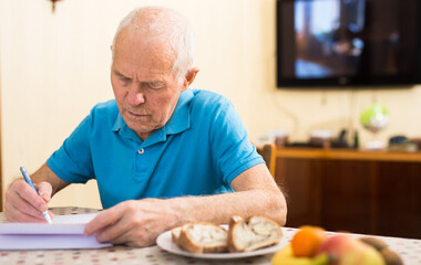 Elderly man writes a letter on a sheet of paper while sitting at a table in room