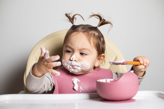 Small Cute Kid Brunette Girl With Two Tails Tasting By Herself With A Spoon And Dirty Hand The Greek Yogurt, Sitting In Baby Highchair With Messy Face And Bib, Self-feeding Concept