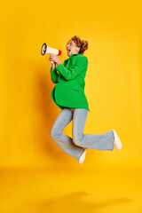 Portrait of young girl in bright green jacket posing, shouting in megaphone in a jump over yellow...