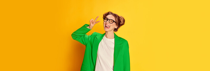 Portrait of young girl in bright green jacket posing with delightful face, grimacing over yellow...