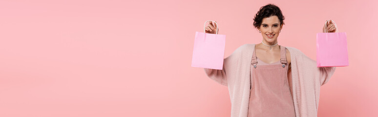 happy brunette woman in warm cardigan showing shopping bags isolated on pink, banner