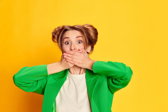 Portrait of young girl in bright green jacket posing, covering mouth with hands over yellow studio background. Secrets