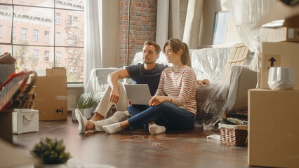 Moving in: Happy Young Homeowners: Couple Sitting on the Floor of their New Apartment Use Laptop...