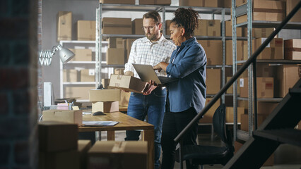 European Male and African Female Working in a Warehouse. Talking, Using Laptop Computer, Checking Retail Stock, Preparing Shipment. Rows of Shelves Full of Cardboard Box Parcels in the Background.
