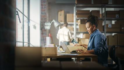 Black Female Warehouse Inventory Manager Works on Laptop Computer while Sitting at Her Desk. In the Background Employee Working in the Room Full of Cardboard Box Parcels Ready For Shipping.