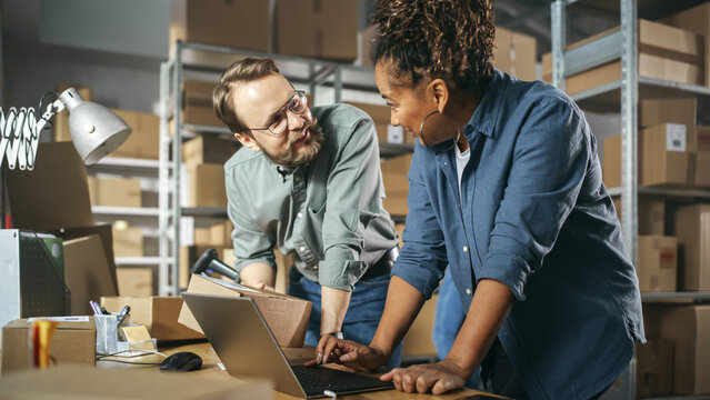 Happy Male and Female Working in Warehouse. Talking, Using Laptop Computer, Checking Retail Stock, Preparing Shipment. Successful Small Business Owners High Five Each Other.