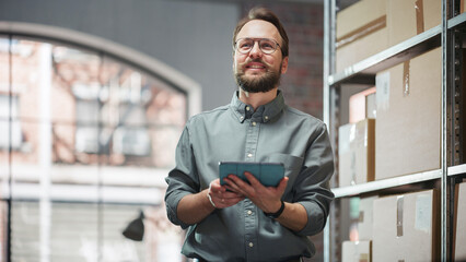 Portrait of a Happy Small Warehouse Employee Checking Inventory, Using Tablet. Handsome Man with Moustache Smiling in a Storeroom with Parcels and Online Purchased Orders Ready for Shipment.