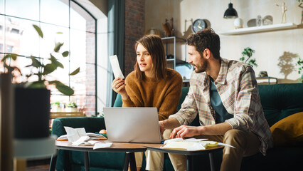 Doing Accounting at Home: Happy Couple Using Laptop Computer, Sitting on Sofa in Apartment. Young...