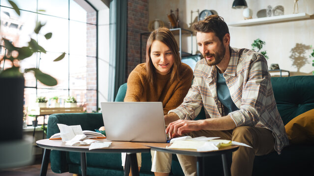 Doing Accounting at Home: Happy Couple Using Laptop Computer, Sitting on Sofa in Apartment. Young Family Filling Tax Forms, Mortgage Documents, Bills, Checks, Balances, Invoices are in Order