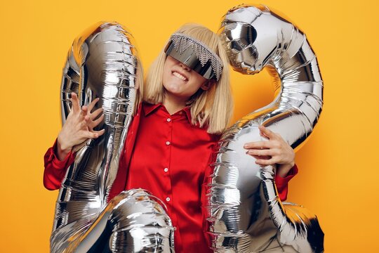 a stylish, fashionable blonde woman stands on a yellow background in futuristic glasses and holds inflatable balloons in the shape of the number twenty-two with her mouth wide open with delight