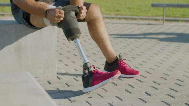 Closeup of male athlete removing his prosthetic leg sitting on parapet outside on sunny day. Man with physical disability taking off artificial leg after working out outdoors. Amputee sport concept.