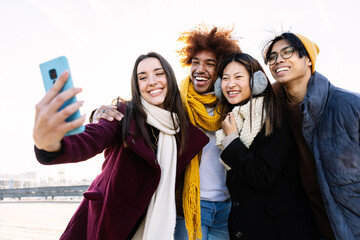 Happy group of teenage friends taking a selfie portrait on winter day. Millennial diverse people on warm clothes enjoying travel vacation together. Friendship lifestyle concept - obrazy, fototapety, plakaty