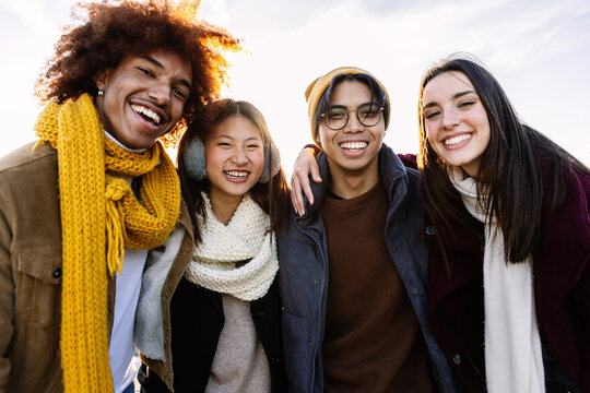 Portrait of four multiracial student friends in winter clothes smiling at camera. Millennial united diverse people standing together outdoors