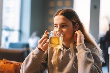 Portrait of a joyful teengirl enjoying a cup of tea at cafe. Smiling beautiful girl drinks hot tea in winter. Teengirl drinking hot tea. Cafe city lifestyle. Colorful portrait of happy pretty girl