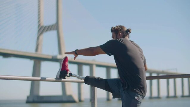 Closeup of strong man with prosthesis doing stretching exercise outdoors. Male athlete with disability putting artificial leg on railing, bending and touching toes. Amputee sport, strength concept.
